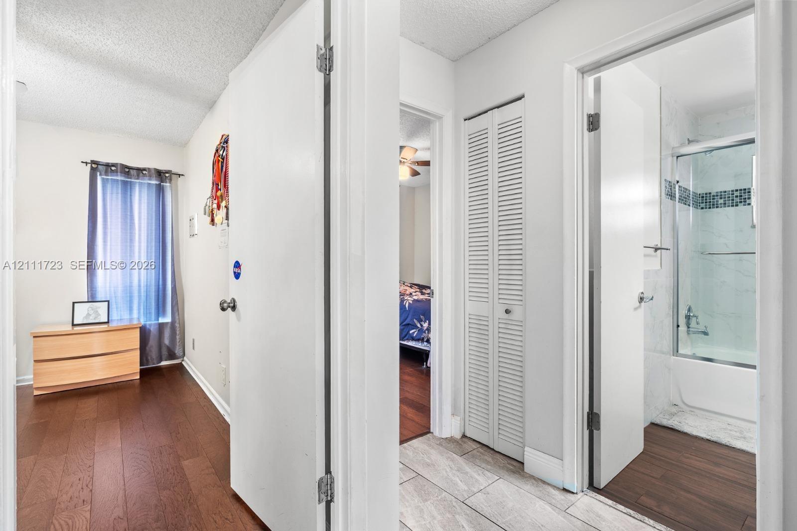 Hallway featuring wood-finish flooring, white louvered closet doors, and tile flooring with access to a bathroom containing a shower-tub combination with tiled surround