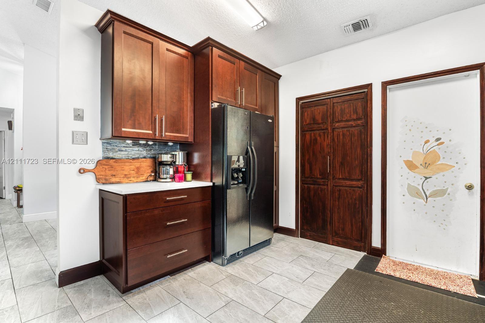 Rich-toned wood cabinetry with bar-style hardware, stainless steel refrigerator, tiled backsplash, and stone-finish flooring