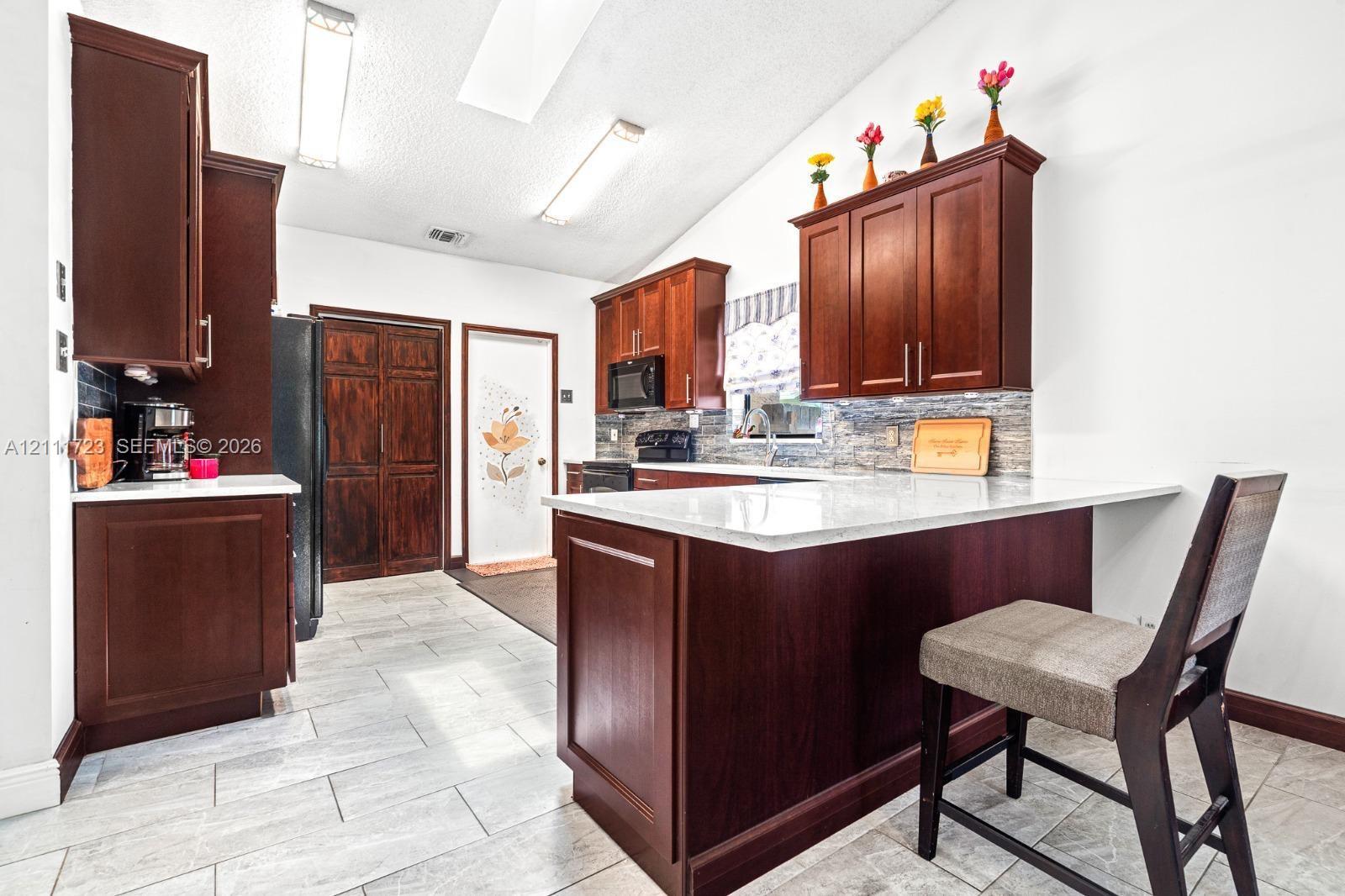 Kitchen featuring dark wood cabinetry, light stone-finish countertops, tile backsplash, tile flooring, and a breakfast bar