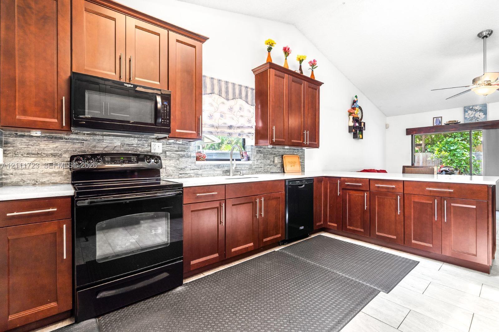 Kitchen featuring wood cabinetry, solid surface countertops, stacked-stone backsplash, black built-in appliances, and vaulted ceilings