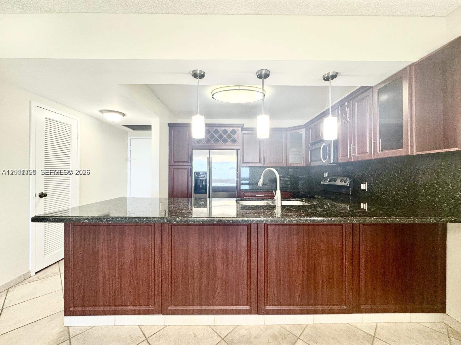 Kitchen featuring dark wood-finish cabinetry, built-in wine rack, stone-finish countertops, three pendant lights, and tile flooring Kitchen featuring dark wood-finish cabinetry, built-in wine rack, stone-finish countertops, three pendant lights, and tile flooring