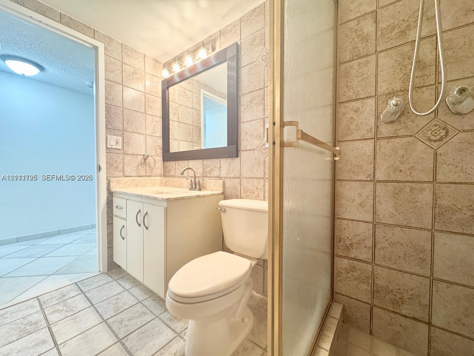 Bathroom featuring a tiled walk-in shower with a gold-tone frame, vanity with neutral-toned countertop, framed wall mirror, and integrated lighting