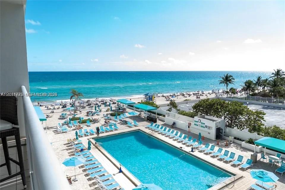 Elevated balcony overlooking an expansive swimming pool, paved pool deck, sandy beach, and ocean shoreline