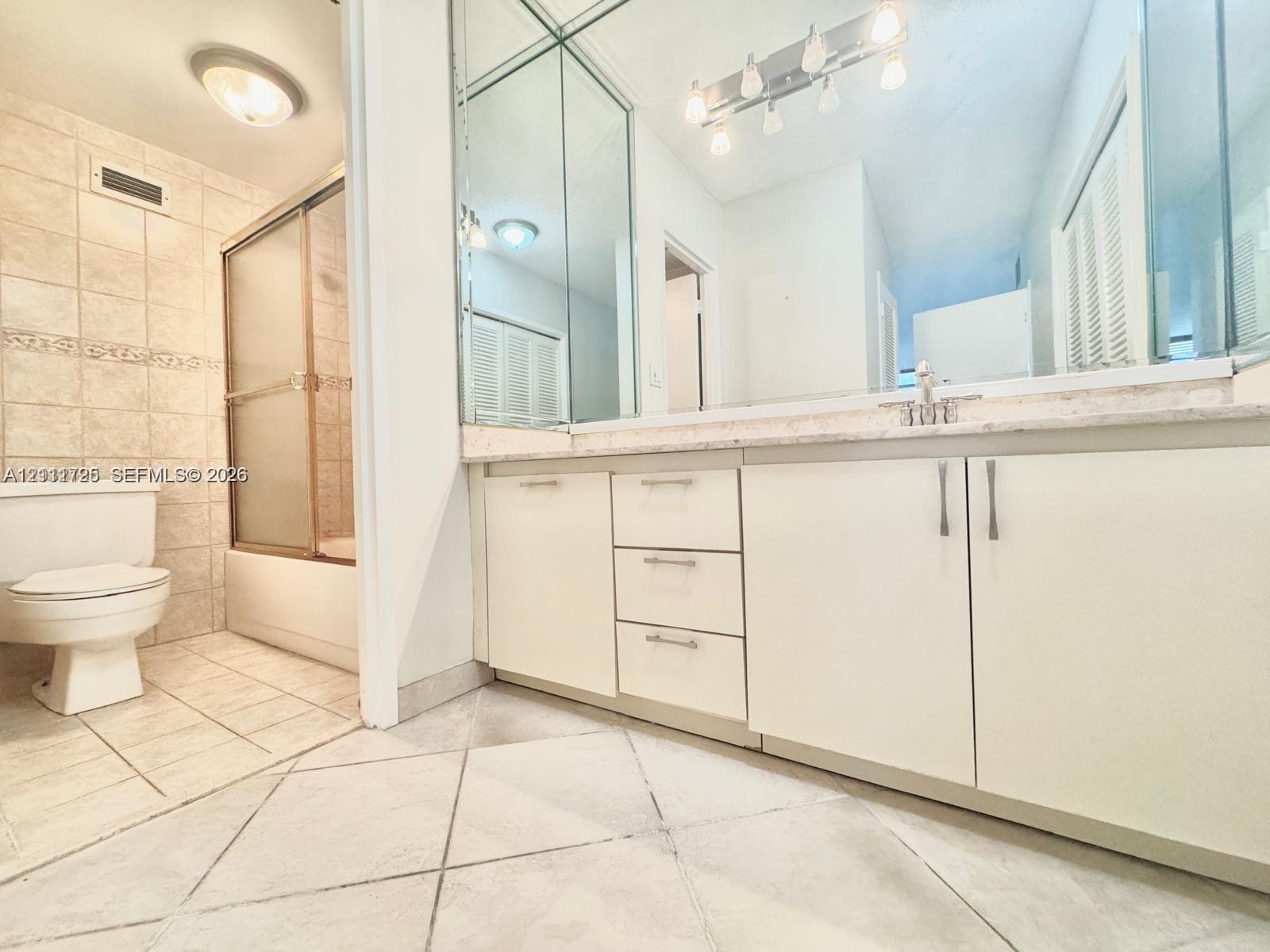 Bathroom featuring a vanity with slab countertop, large-format tiled flooring, full-wall mirror, shower enclosure with tiled surround, and cabinetry with metal hardware