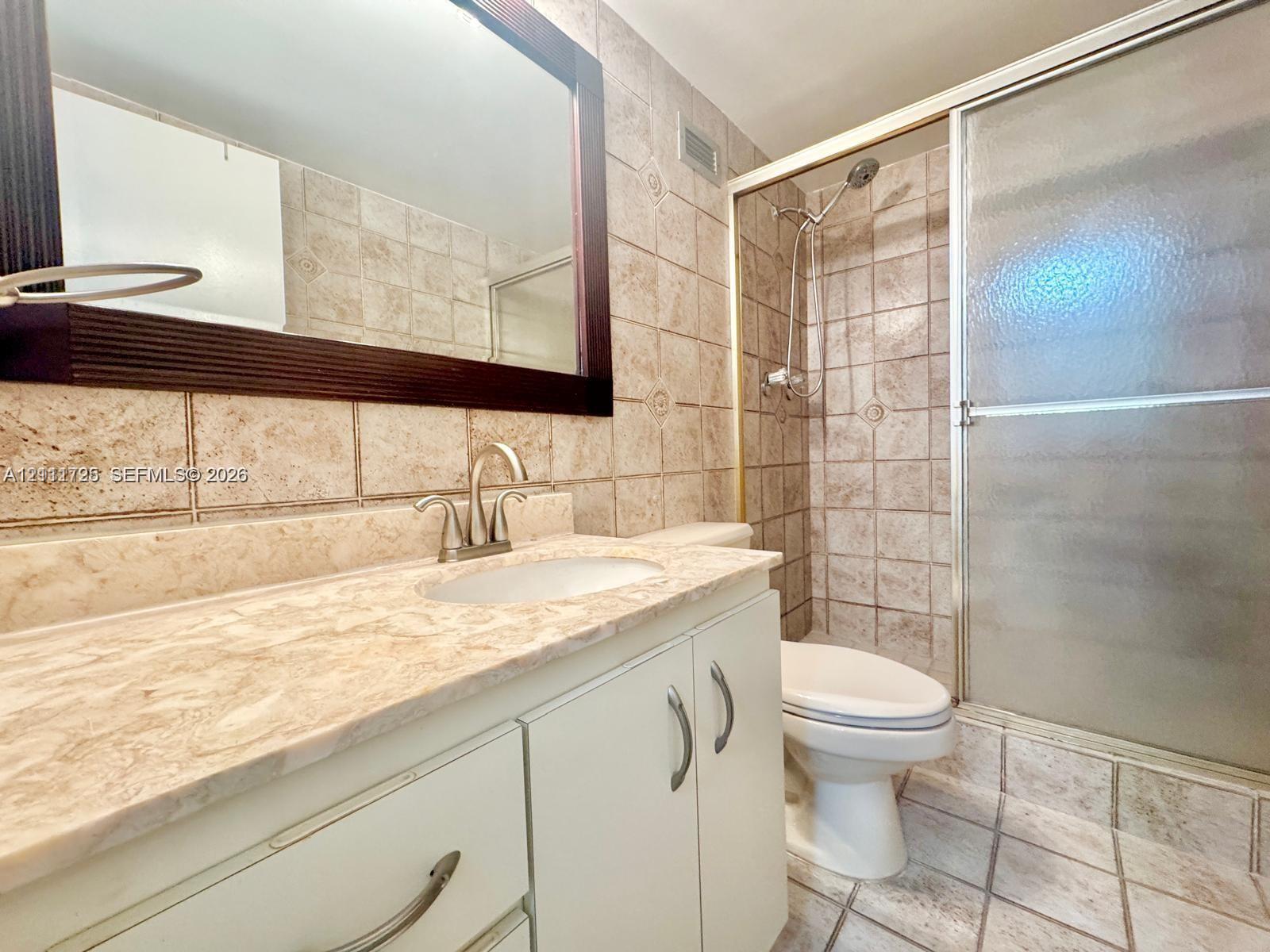 Bathroom featuring a vanity with stone-finish countertop, undermount sink, metal hardware, framed mirror, and a walk-in shower enclosure with tile surround