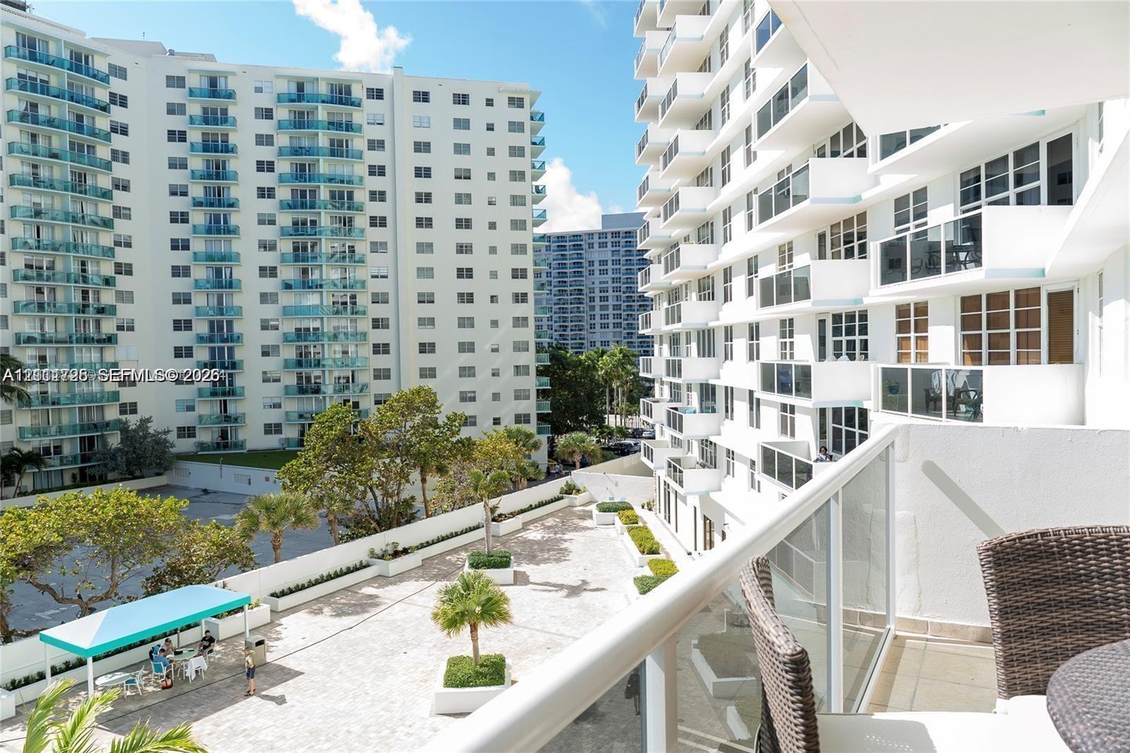 Private balcony with glass panel railing overlooking a paved outdoor terrace featuring mature palms, raised planters, and a shaded seating area