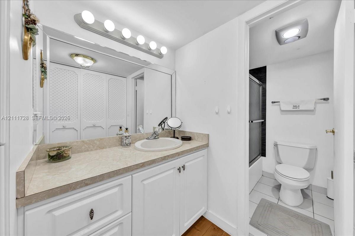 Bathroom featuring white cabinetry, solid-surface countertop with an integrated basin, large wall-mounted mirror, overhead multi-bulb lighting, and a tub-shower enclosure with tiled surround