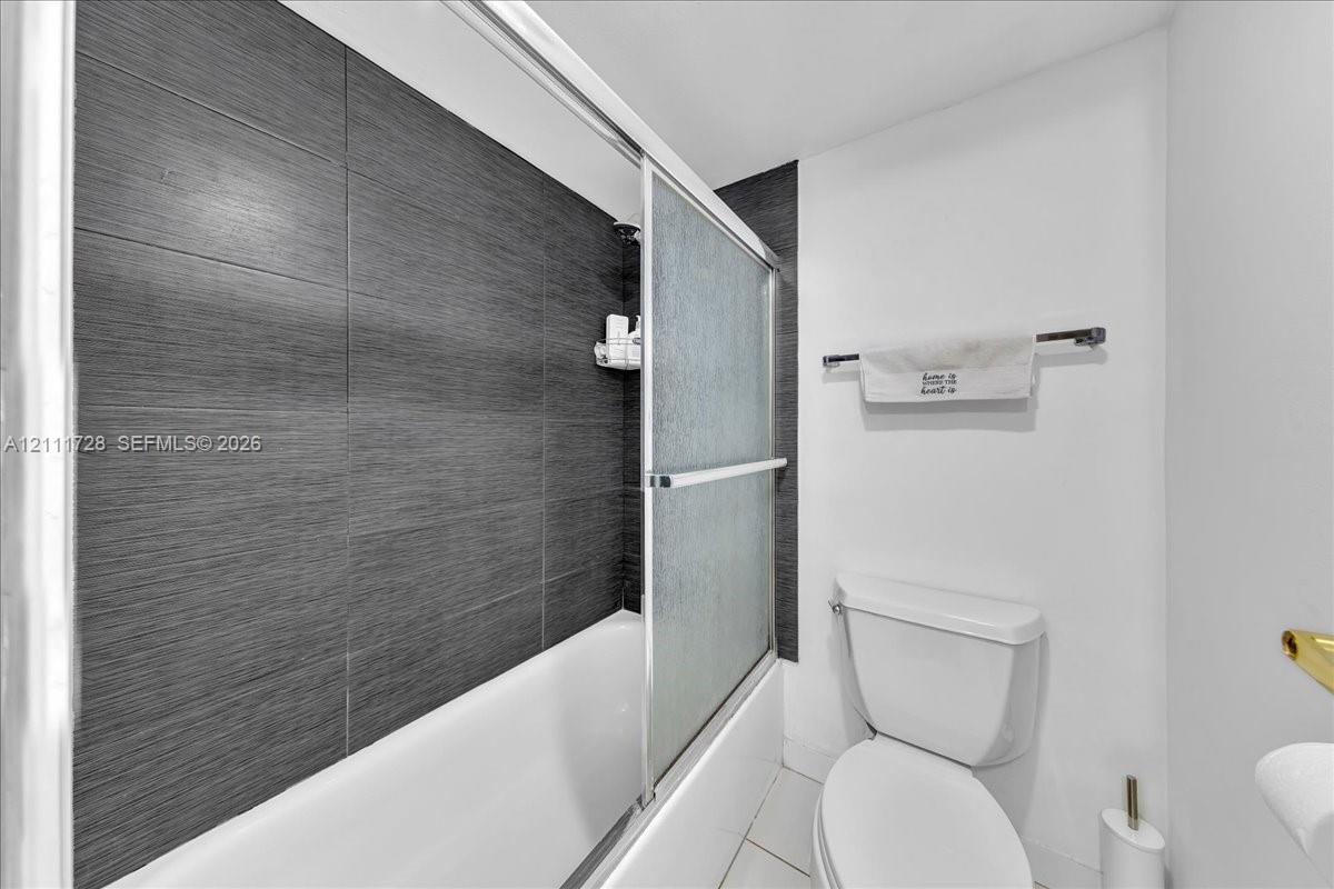 Bathroom featuring large-format dark grey textured tile shower surround, sliding glass shower door, white acrylic tub, and elongated porcelain toilet
