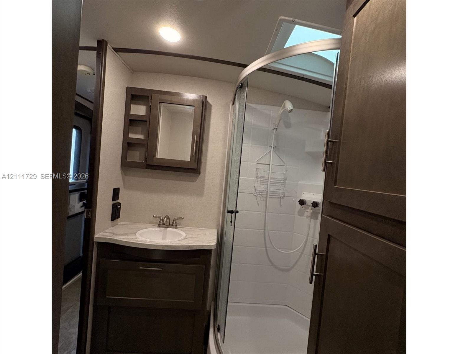Bathroom featuring dark wood-finish cabinetry, a stone-pattern vanity countertop with an integrated basin, a curved glass-enclosed shower, a skylight, and wall-mounted medicine cabinet Bathroom featuring dark wood-finish cabinetry, a stone-pattern vanity countertop with an integrated basin, a curved glass-enclosed shower, a skylight, and wall-mounted medicine cabinet