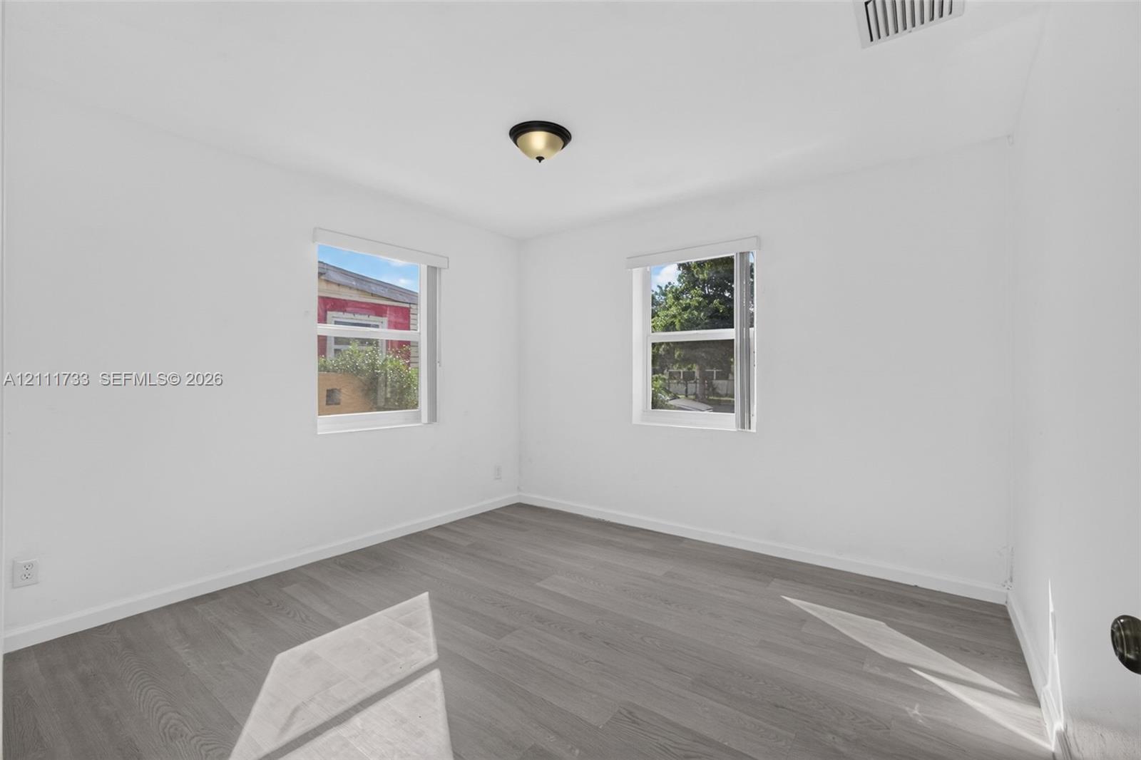 Larger Front bedroom - dual exterior view windows featuring white walls, wood-finish flooring, and windows with horizontal blinds, illuminated by a ceiling-mounted flush light fixture