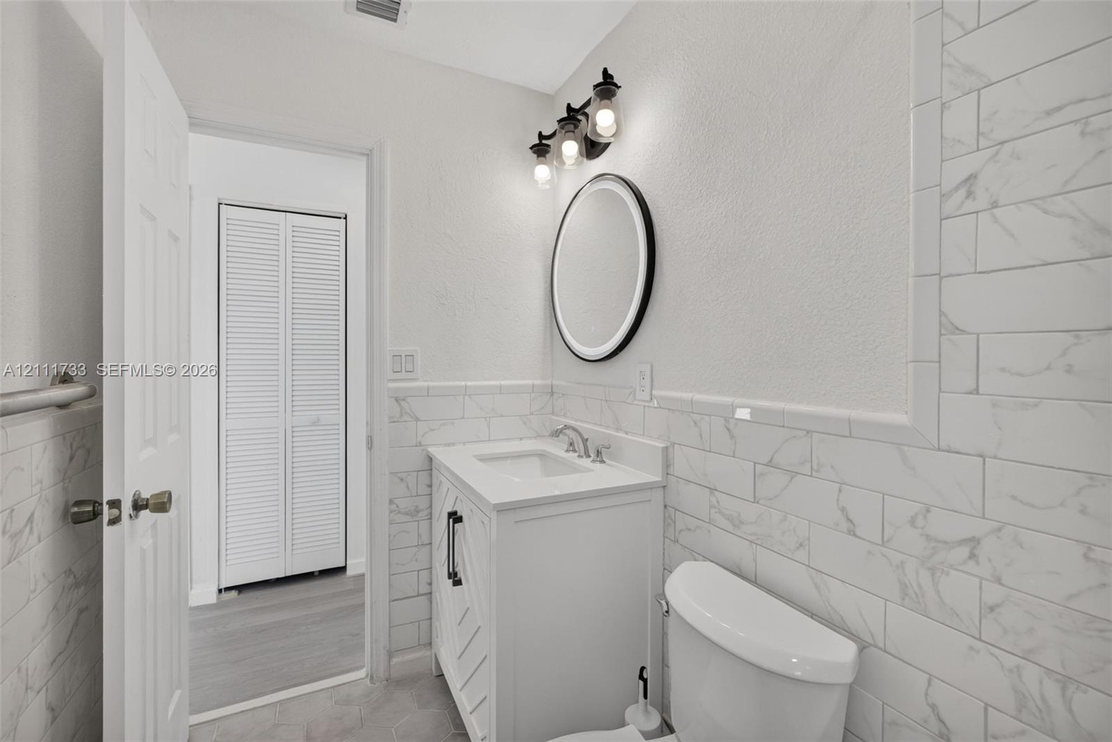 Main Bathroom featuring a white vanity cabinet with chevron detailing, a round backlit wall mirror, stone-finish subway wall tiles, and a three-light fixture