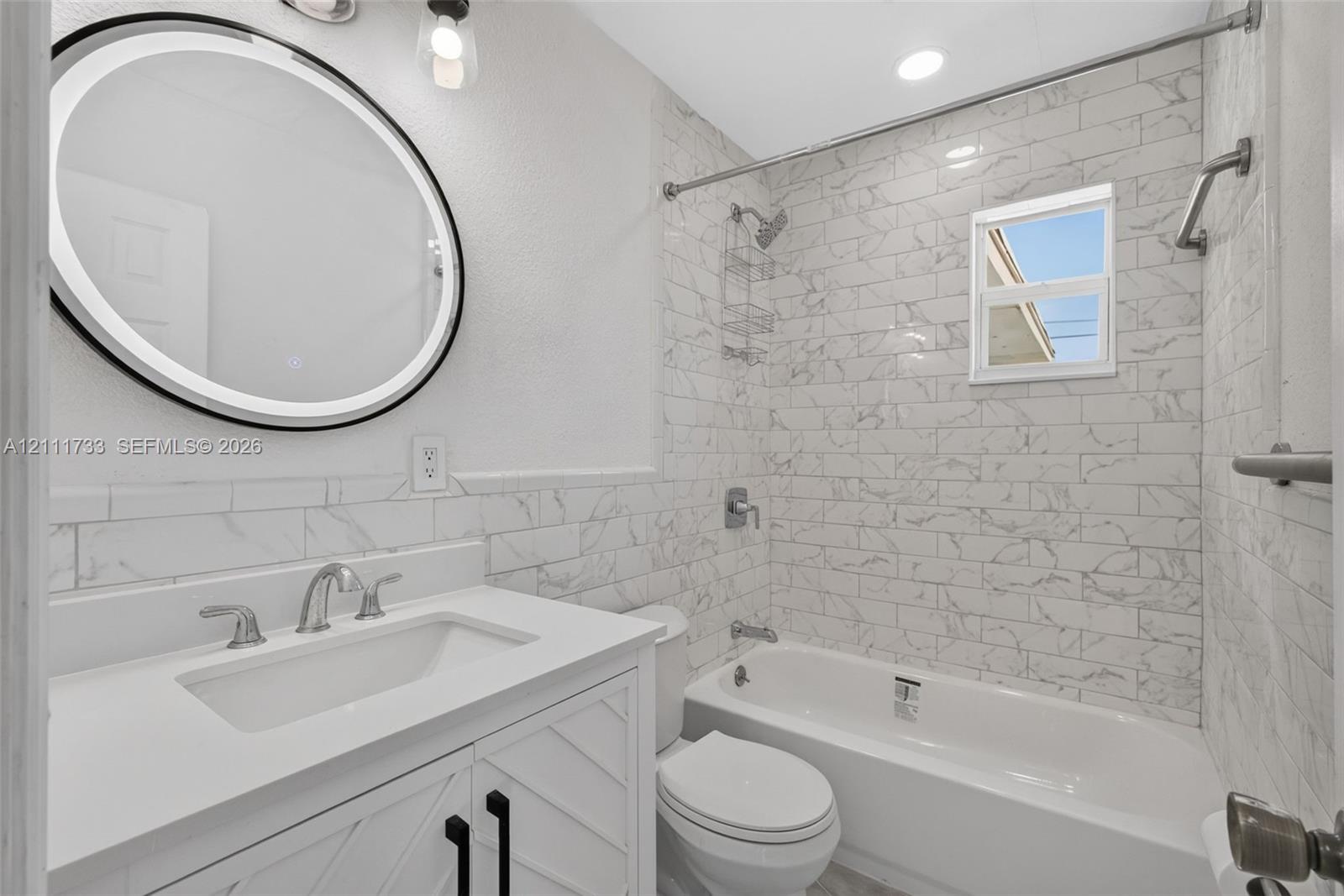 Main Bathroom featuring a white vanity with geometric cabinet detailing, a quartz countertop, an integrated vessel sink, a circular LED-backlit mirror, and white subway-patterned tile wall surrounds with a marble-style finish