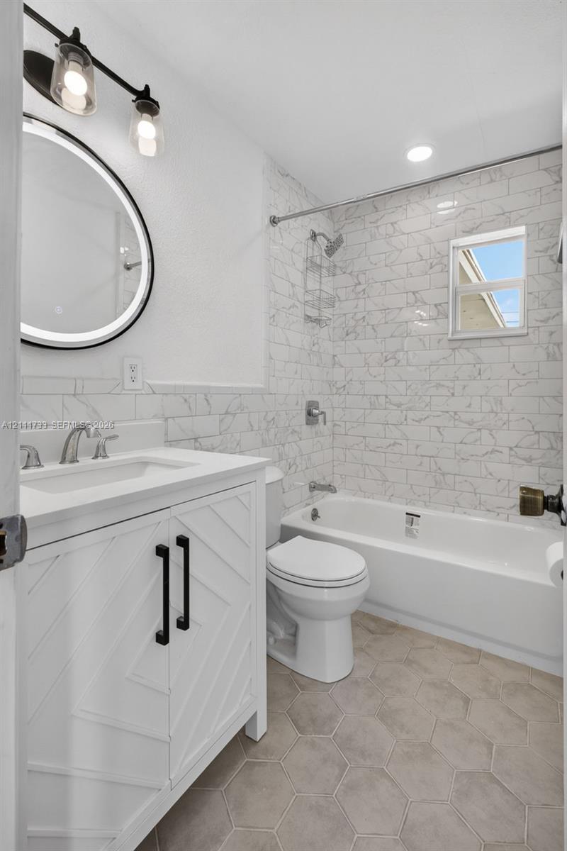 Main Bathroom featuring a white chevron-pattern vanity with a solid-surface countertop, an illuminated round mirror, porcelain tile shower surround, and hexagonal floor tile