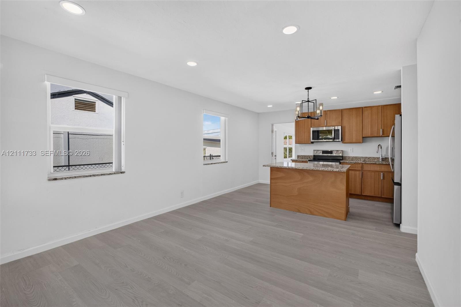 Open-concept kitchen and living area featuring wood-finish flooring, recessed lighting, medium-tone wood cabinetry, stone-surfaced countertops, and a central island