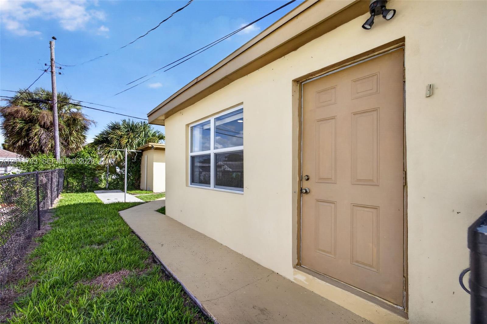 Exterior side elevation featuring a smooth-stucco finish, six-panel entry door, fixed-frame window, concrete walkway, and exterior floodlighting. Outside entry to Rear Bedroom.