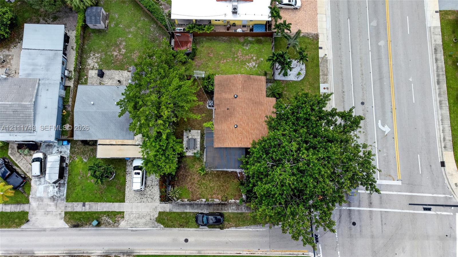 Aerial perspective of residential property featuring a shingle roof, mature landscaping, paved driveway, and grass lawn area