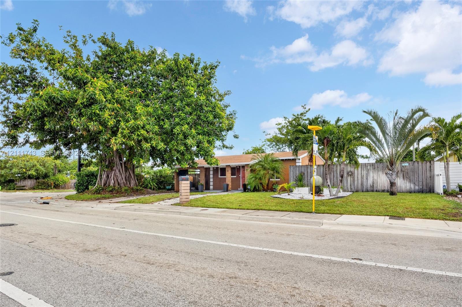 Single-story residence featuring a horizontal wood-slat privacy fence, manicured front lawn, mature specimen tree, and tropical landscaping