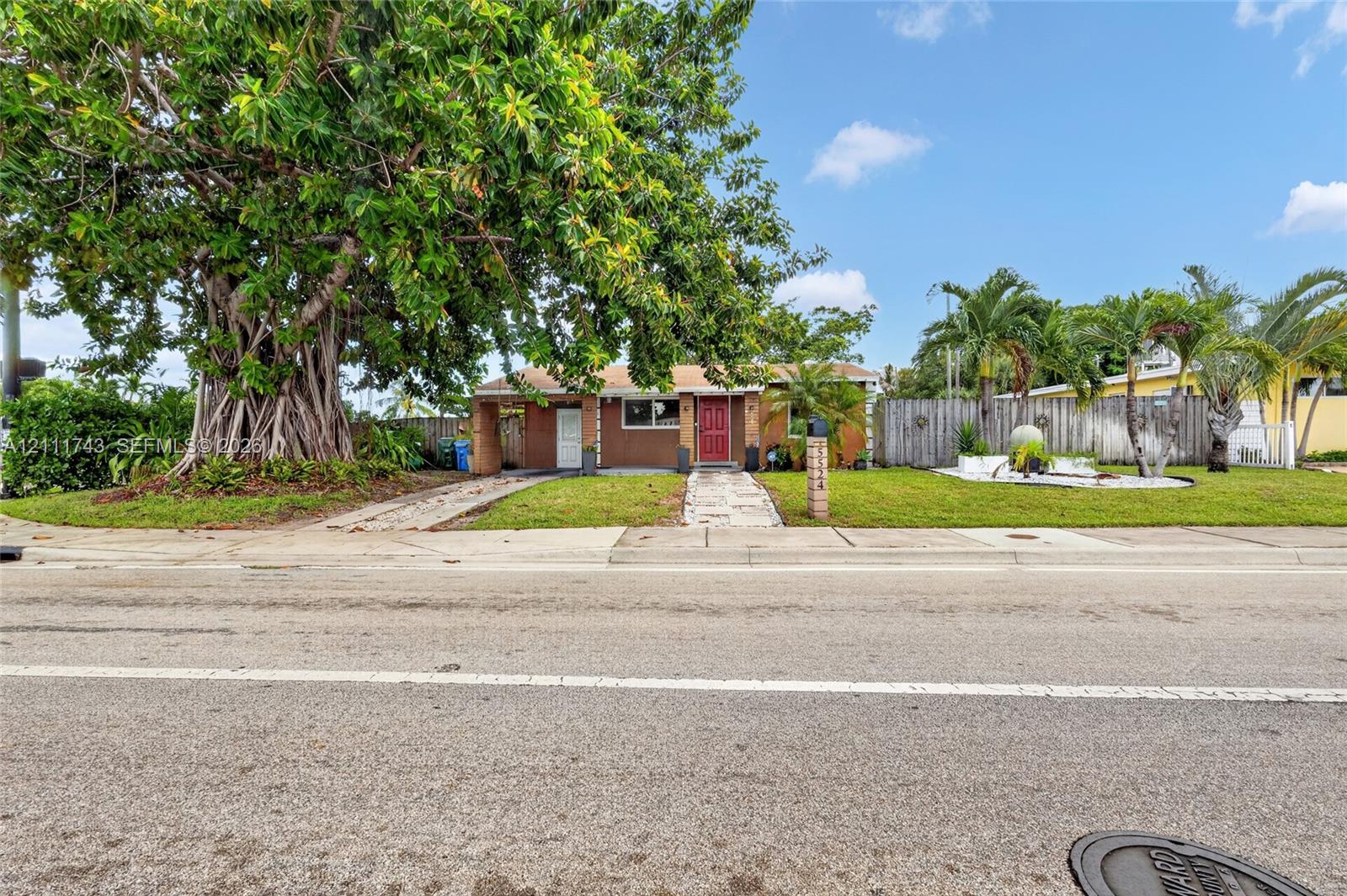 Single-story residence featuring a masonry exterior, red entry door, mature canopy tree, manicured lawn, and wood-style privacy fencing