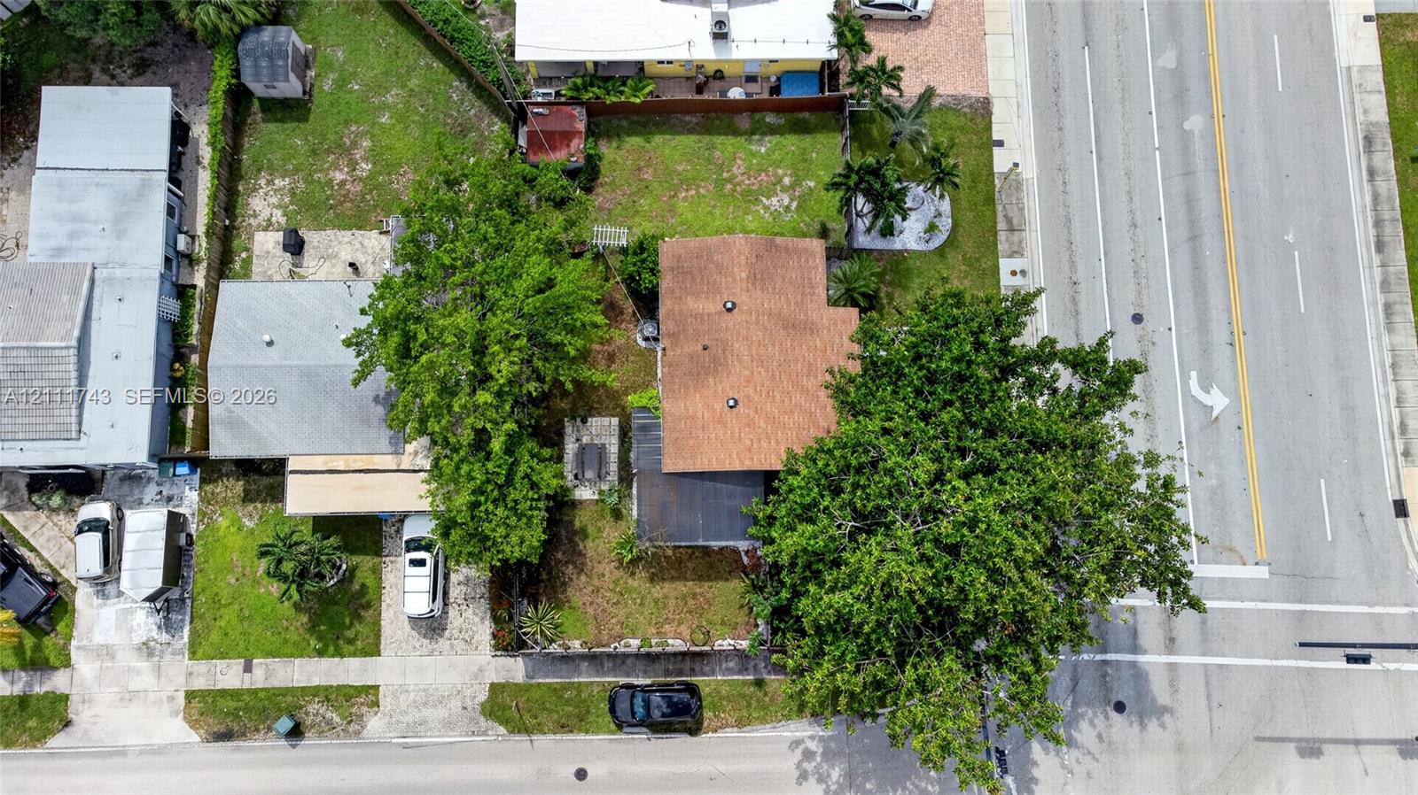 Aerial perspective of the property featuring a structured lot, paved driveway, mature trees, and designated hardscaped area