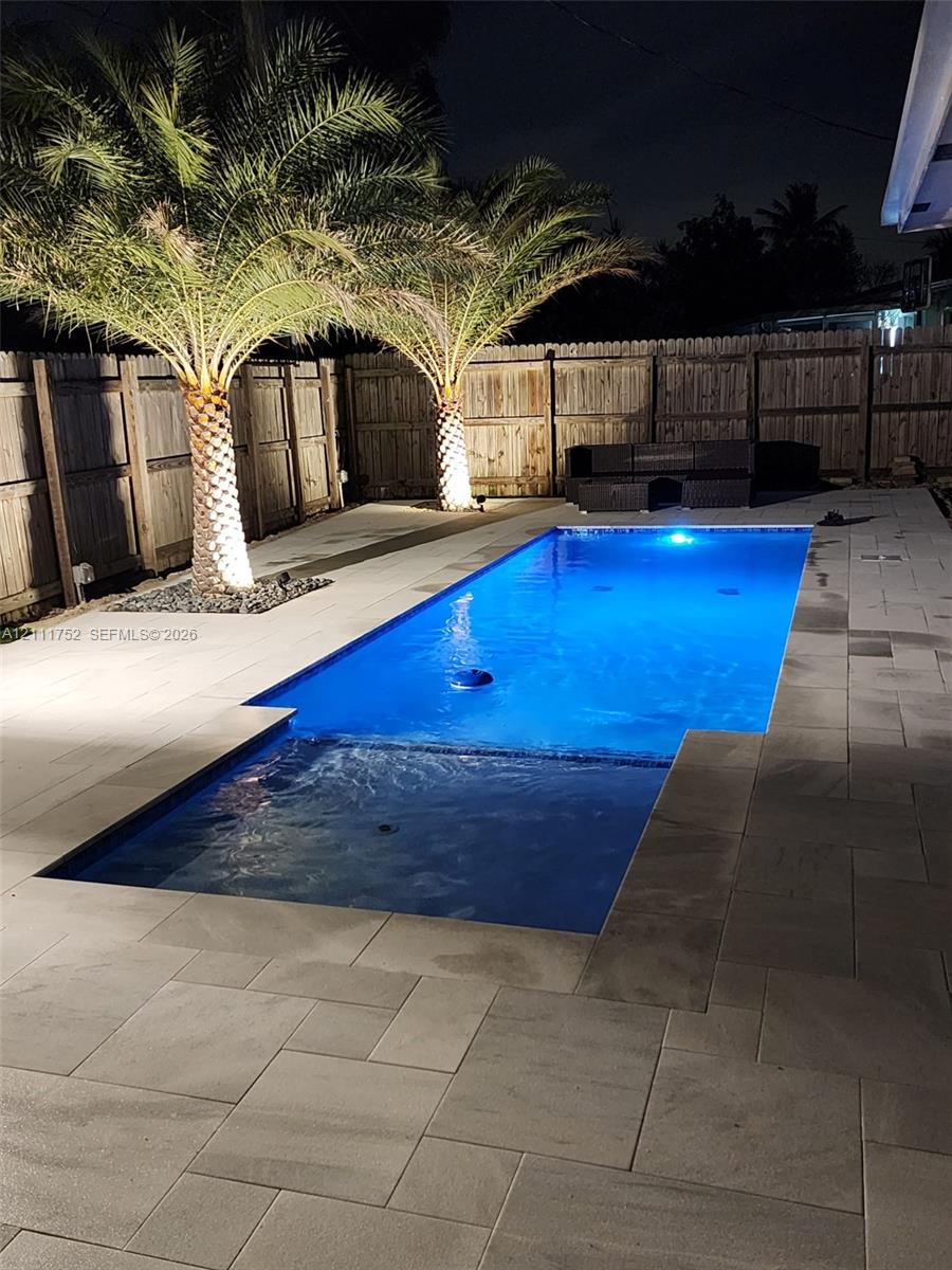 Rectangular pool featuring a shallow entry shelf, expansive stone paver decking, mature palm trees, and perimeter wood privacy fencing