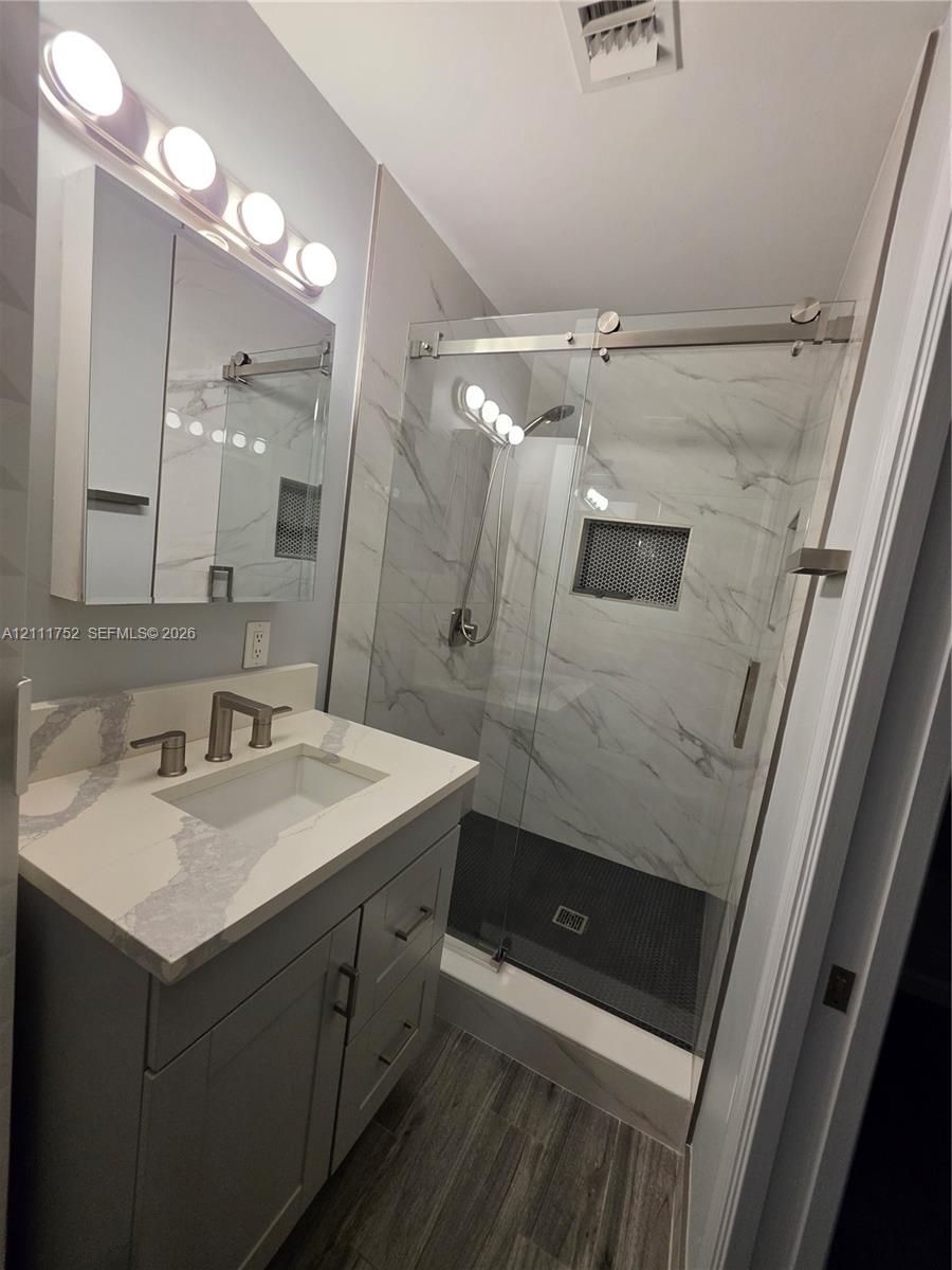 Bathroom vanity featuring a light-toned countertop, rectangular undermount sink, and brushed-finish fixtures, paired with a glass-enclosed walk-in shower with stone-patterned wall tile, a mosaic-tiled recessed niche, and wood-finish flooring