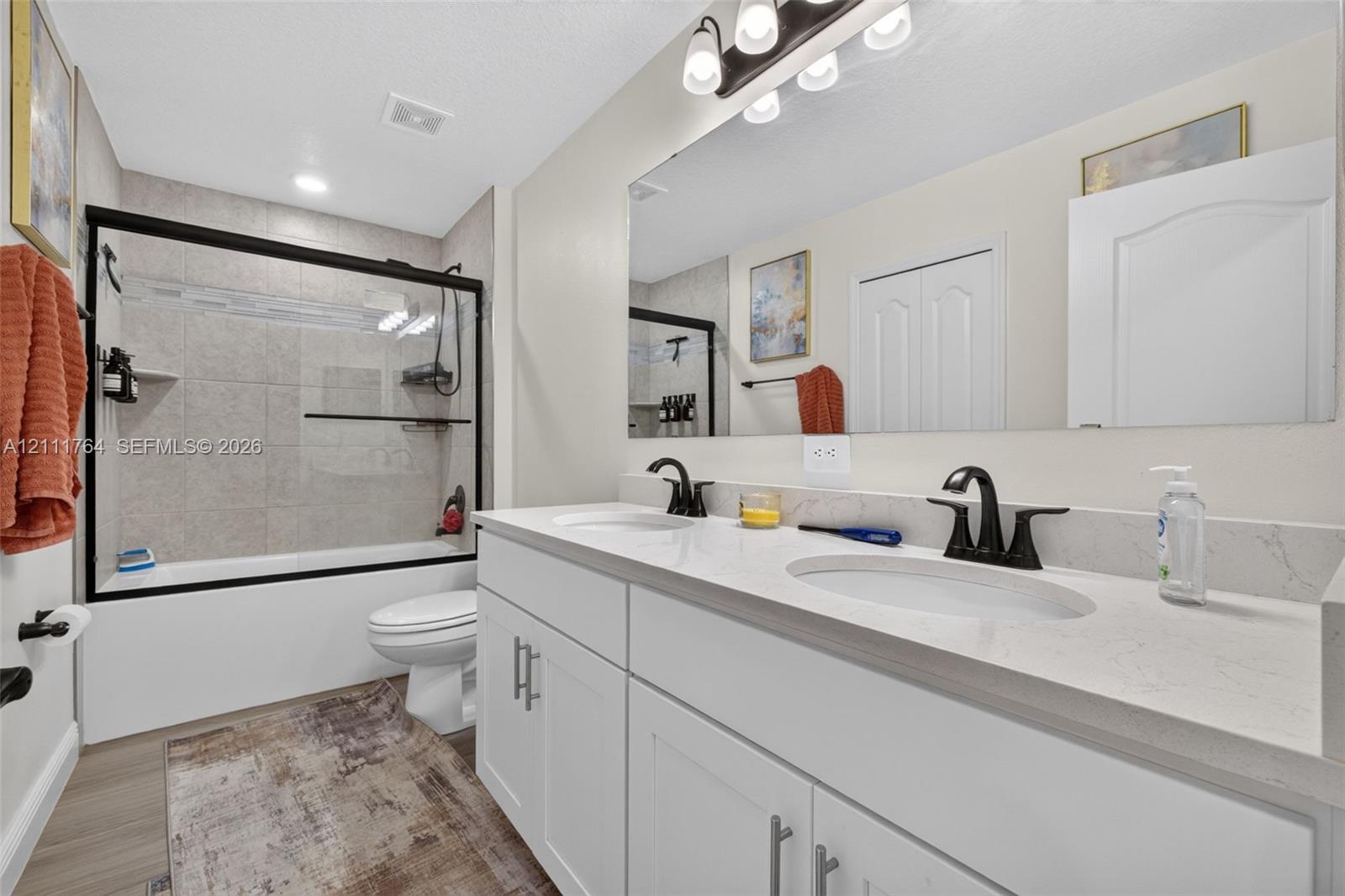 Dual-sink vanity with white cabinetry, light-toned stone-finish countertop, and matte black hardware, paired with a glass-enclosed shower featuring neutral tile surround and matte black fixtures