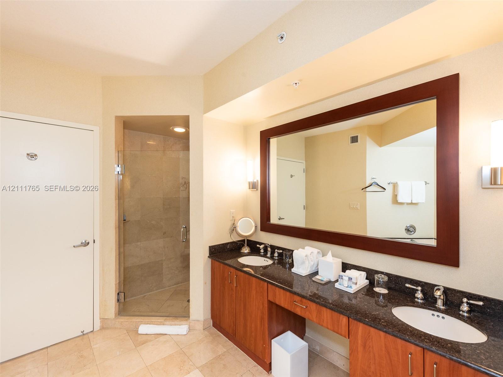 Dual-sink vanity with dark stone countertops, wood-finish cabinetry, a large framed wall mirror, and a walk-in shower with stone tile surround