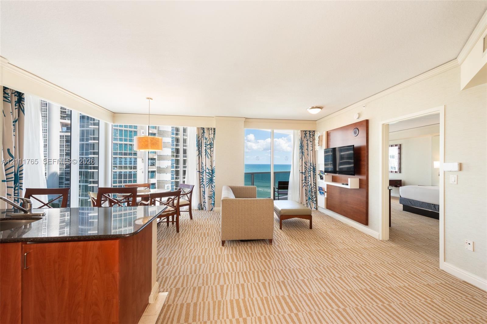 Open-concept living space featuring floor-to-ceiling windows, built-in wooden media cabinetry, a kitchen island with stone countertop, and a private balcony with water scenery