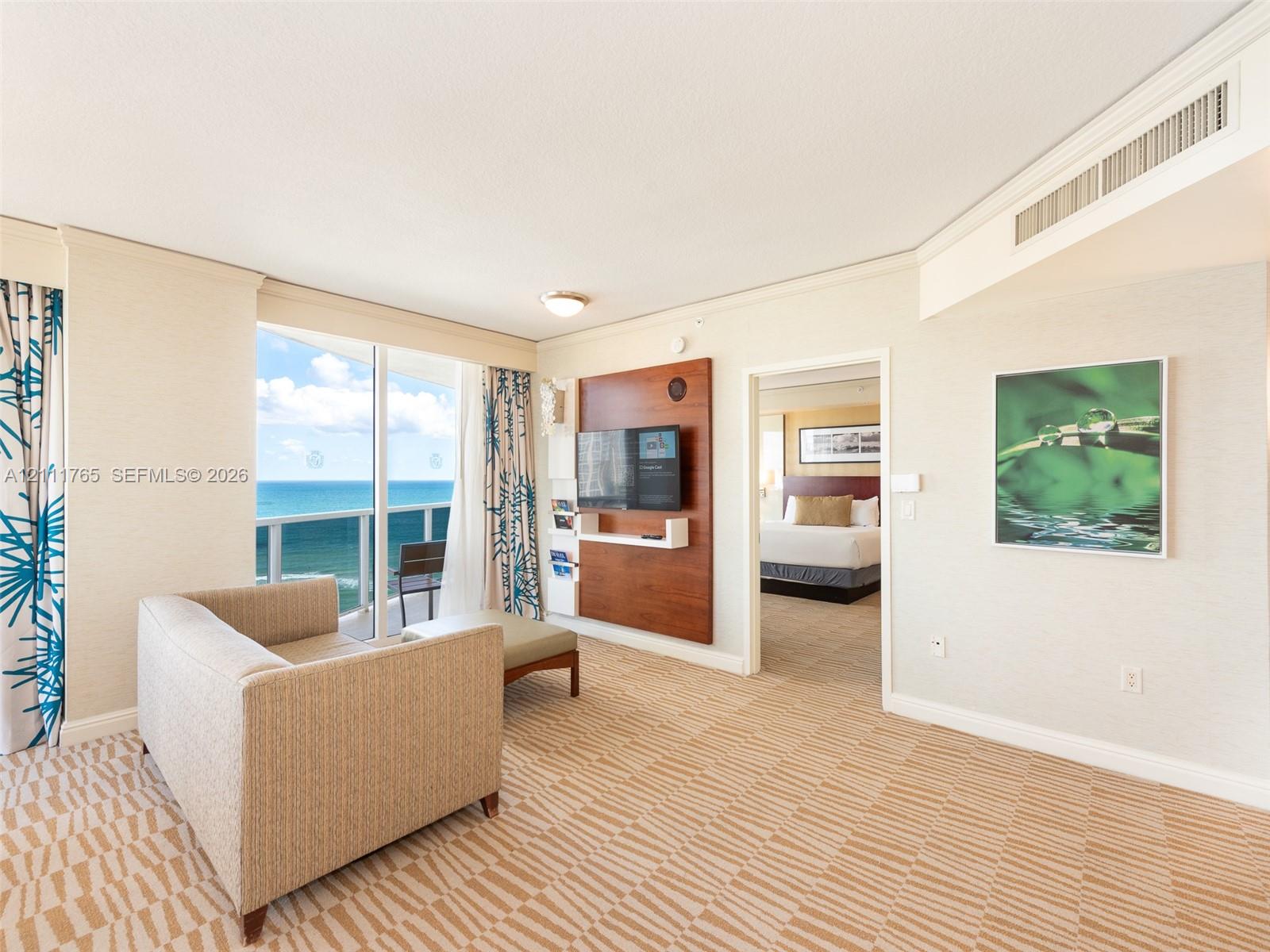 Sliding glass door opening to a private balcony with ocean scenery, wall-mounted wood-finish media console, and patterned wall-to-wall carpeting