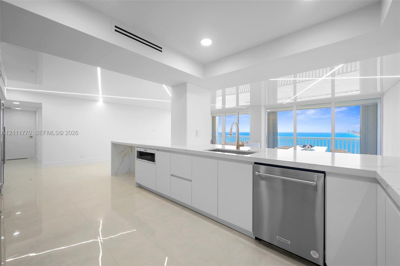 Minimalist kitchen featuring matte white flat-panel cabinetry, light-toned stone-finish countertops, recessed linear lighting, large-format polished tile flooring, and floor-to-ceiling windows with coastal scenery