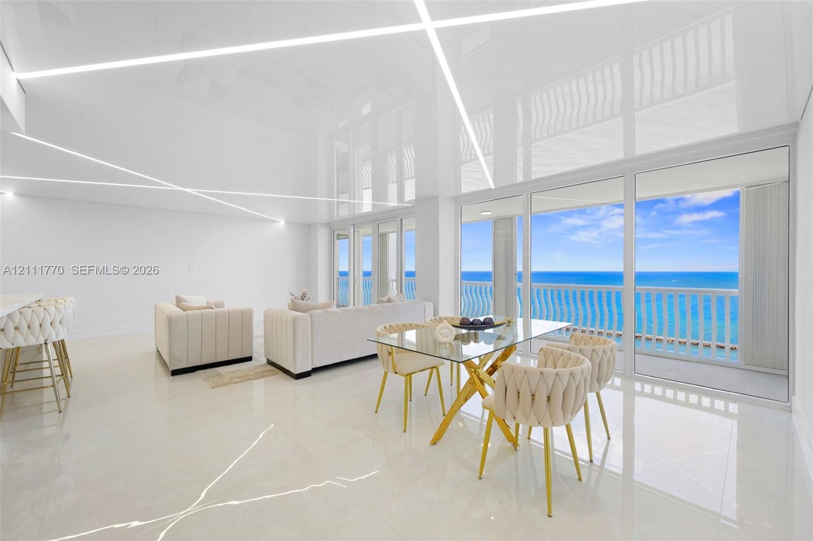 High-gloss reflective ceiling with integrated linear lighting, large-format tile flooring, floor-to-ceiling glass sliding doors, and private balcony with ocean backdrop