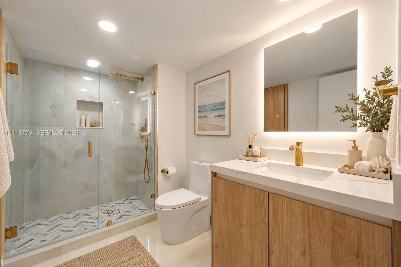 Bathroom featuring a frameless glass walk-in shower with stone tile surround, chevron-patterned floor tile, wood-finish vanity with a solid surface countertop, and a back-lit wall mirror
