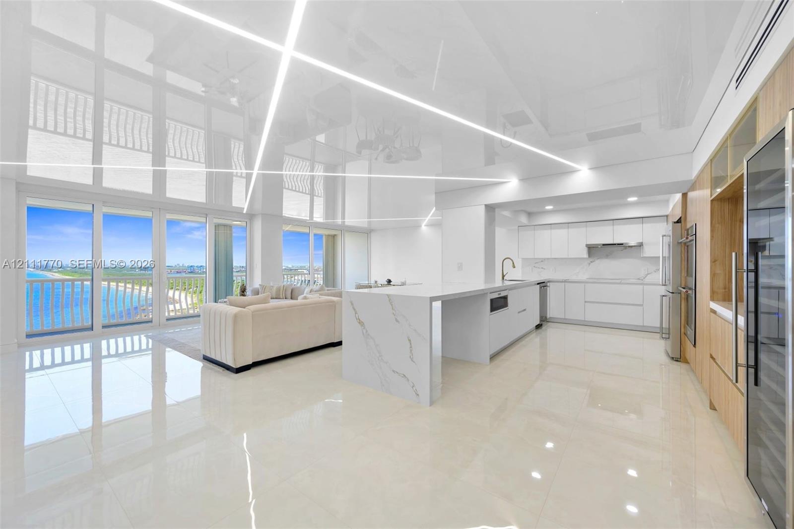 Open-concept living space featuring high-gloss reflective ceiling panels with linear LED lighting, floor-to-ceiling windows, polished stone-finish flooring, and a kitchen island with stone-finish waterfall edges