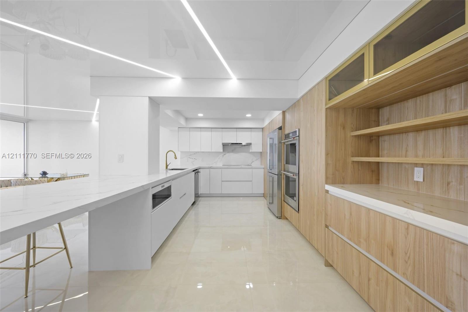 Modern kitchen featuring high-gloss cabinetry, stone-surface countertops, built-in double ovens, linear recessed ceiling lighting, and integrated wood-finish cabinetry