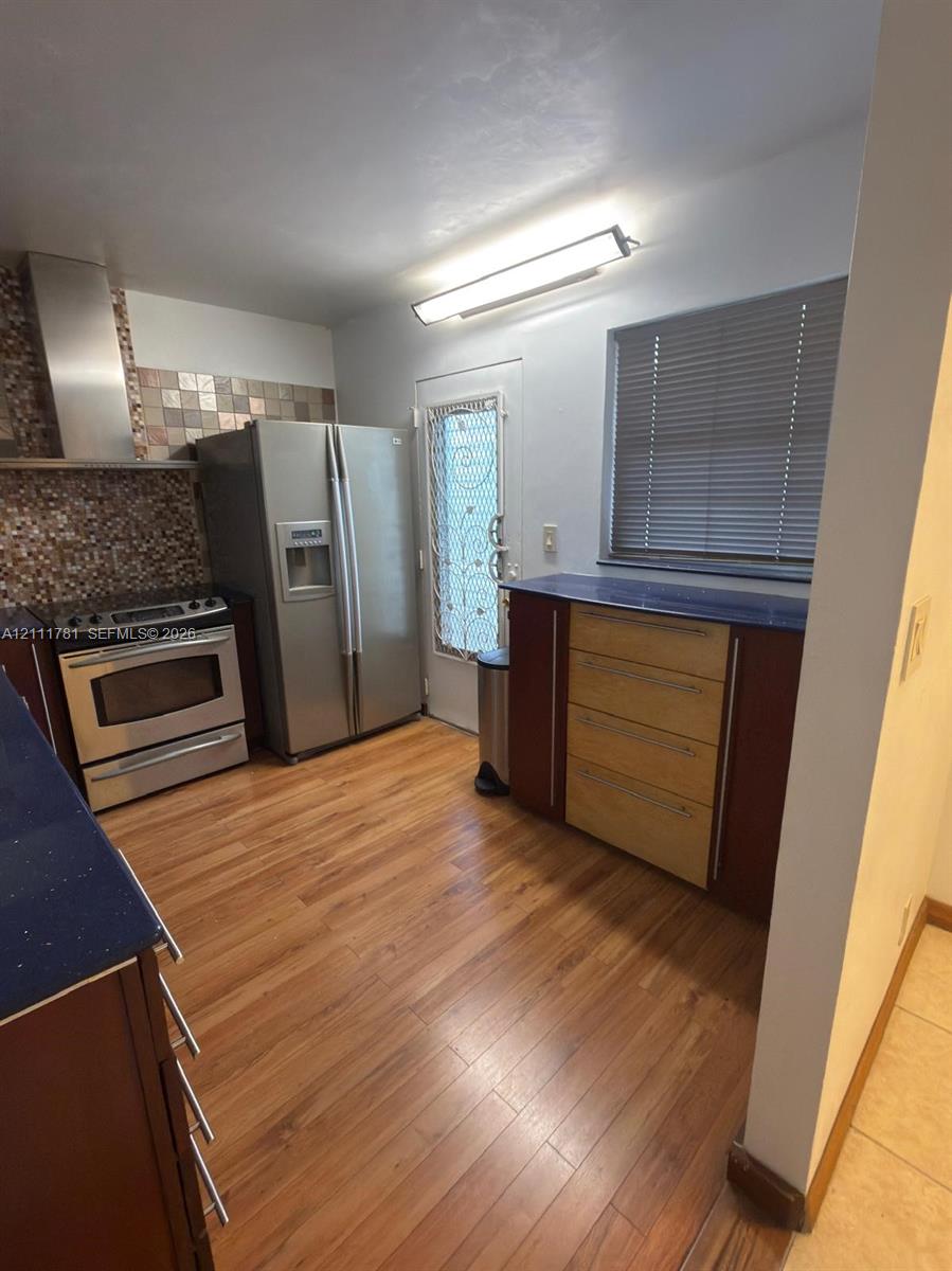 Kitchen featuring wood-finish flooring, stainless steel appliances, mosaic tile backsplash, solid-surface countertops, and wood-toned cabinetry