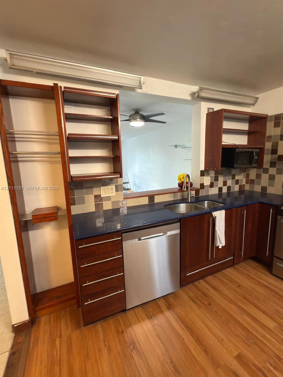 Kitchen featuring wood cabinetry with built-in open shelving, dark countertops, a double-basin stainless steel sink, stainless steel dishwasher, and multi-toned tile backsplash
