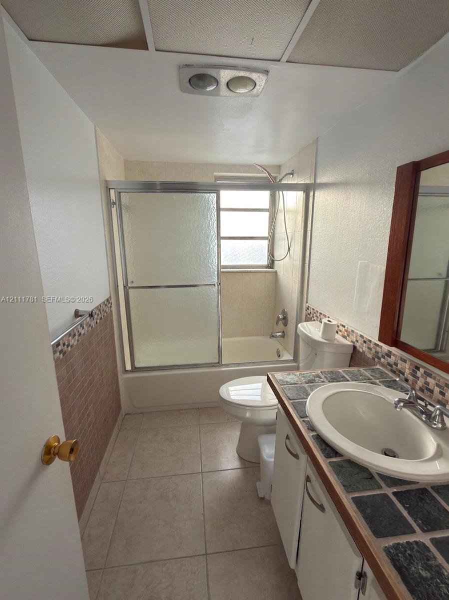 Bathroom featuring a tub-shower combination with sliding glass doors, tile-topped vanity with an integrated basin, tile flooring, and decorative mosaic wall tile accents