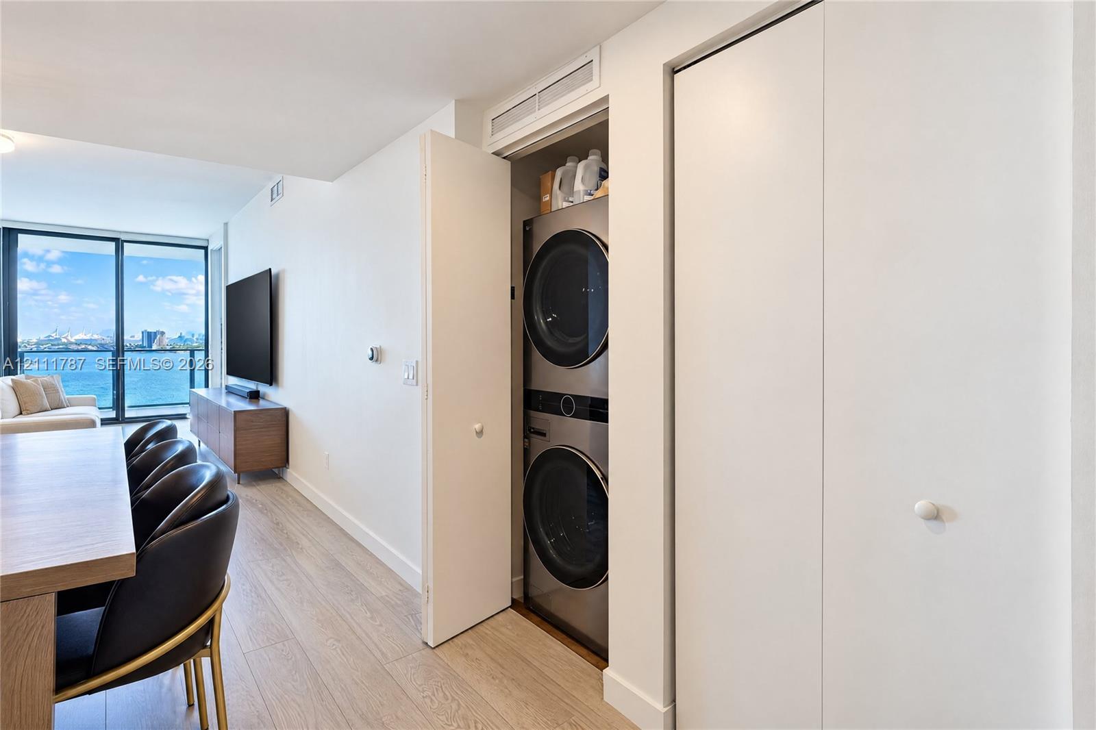 Stacked metallic finish laundry appliances housed in a built-in closet with white doors, featuring wood-finish flooring and floor-to-ceiling glass sliding doors