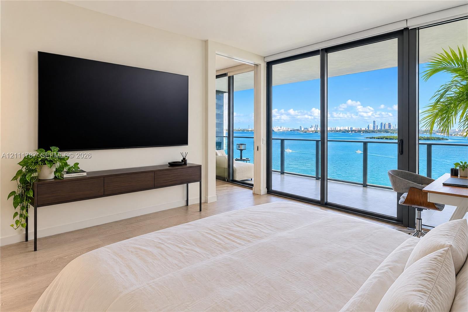 Floor-to-ceiling sliding glass doors, private balcony with metal railing, light-tone wood-finish flooring, and expansive water and skyline backdrop