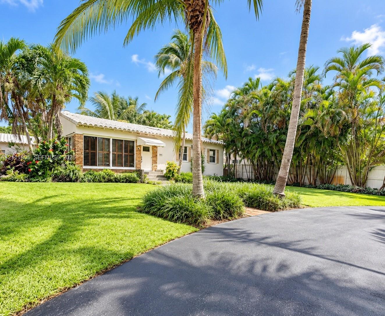 Stucco exterior residence featuring a clay tile roof, stone accent siding, multi-pane windows, and a paved driveway, surrounded by mature palm trees and landscaped grounds