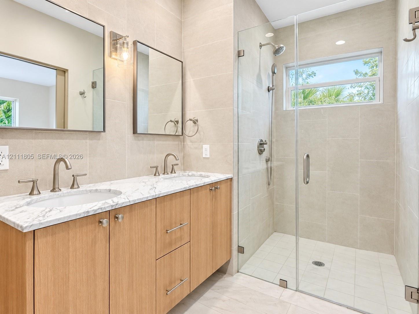 Double vanity featuring natural wood-finish cabinetry, a stone-patterned countertop, dual under-mount sinks, frameless glass-enclosed walk-in shower, and large-format wall tiling