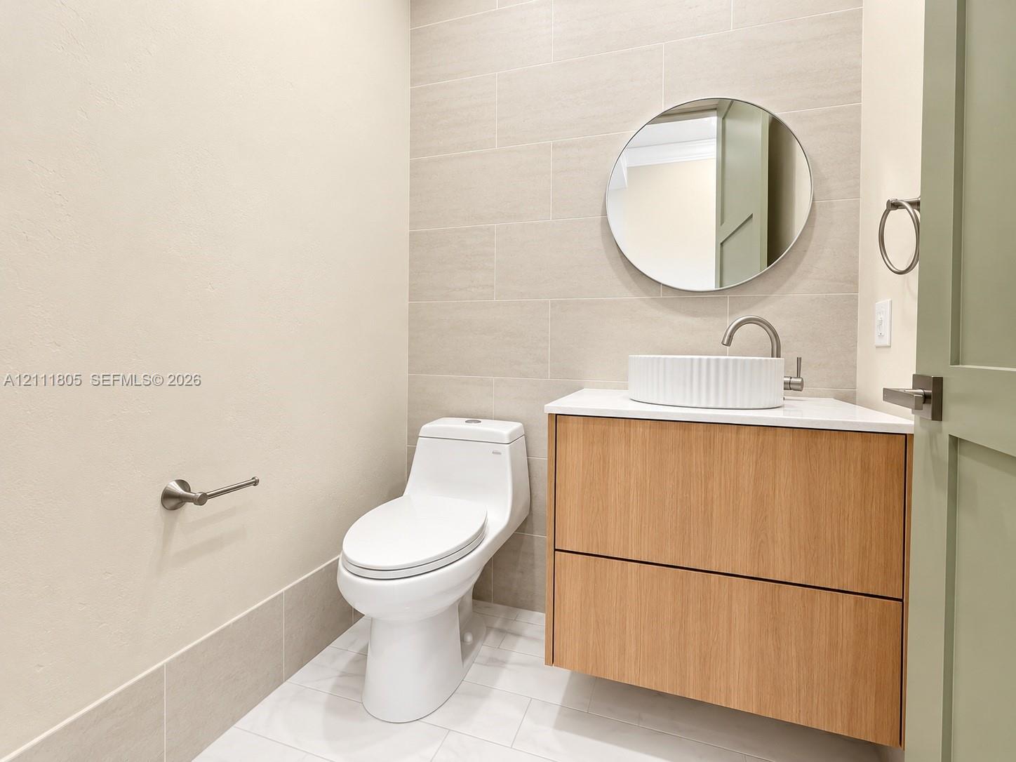 Floating wood-finish vanity with a white vessel sink, neutral rectangular wall tile, round mirror, and one-piece toilet