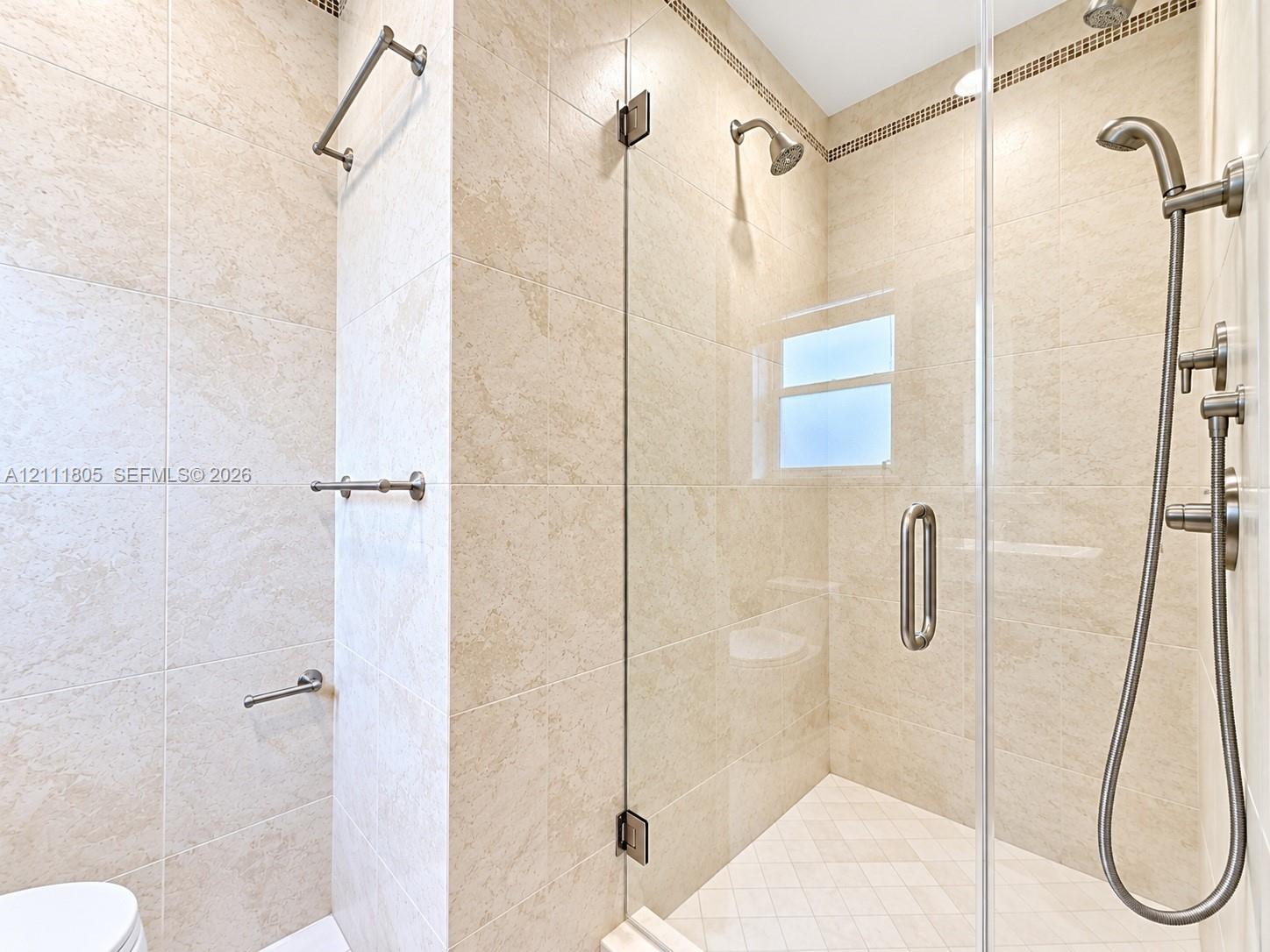 Walk-in shower featuring floor-to-ceiling neutral tile, glass enclosure with metal hardware, rainfall showerhead, handheld shower sprayer, and mosaic tile accent detailing