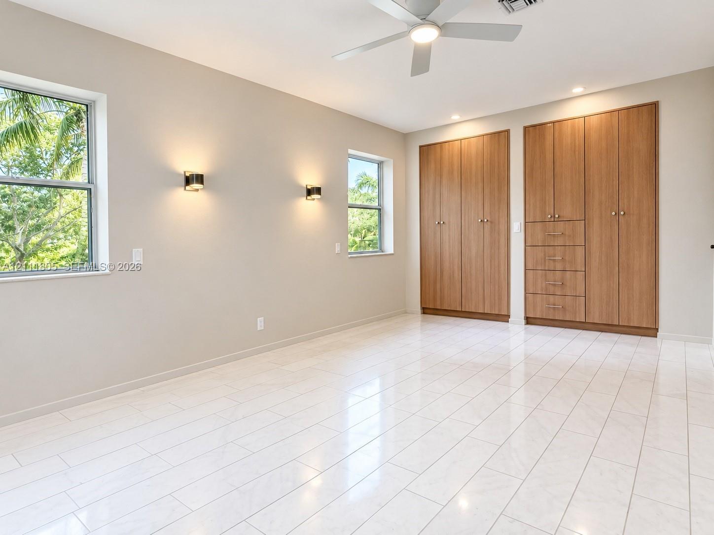 Neutral-toned interior featuring large-format tile flooring, built-in floor-to-ceiling wood-finish cabinetry, wall-mounted light fixtures, and a ceiling fan with integrated lighting