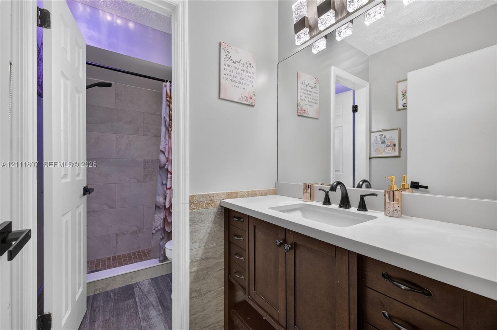 Bathroom featuring a dark wood vanity with white countertop, undermount sink, matte black widespread faucet, large wall-mounted mirror with integrated lighting, and a walk-in shower with grey tile surround