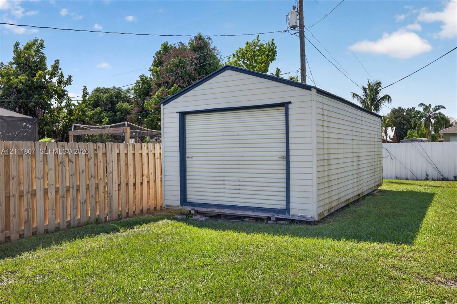 Detached storage structure with metal siding, a roll-up garage door, and a gabled roof, situated on a lawn enclosed by wood and vinyl privacy fencing