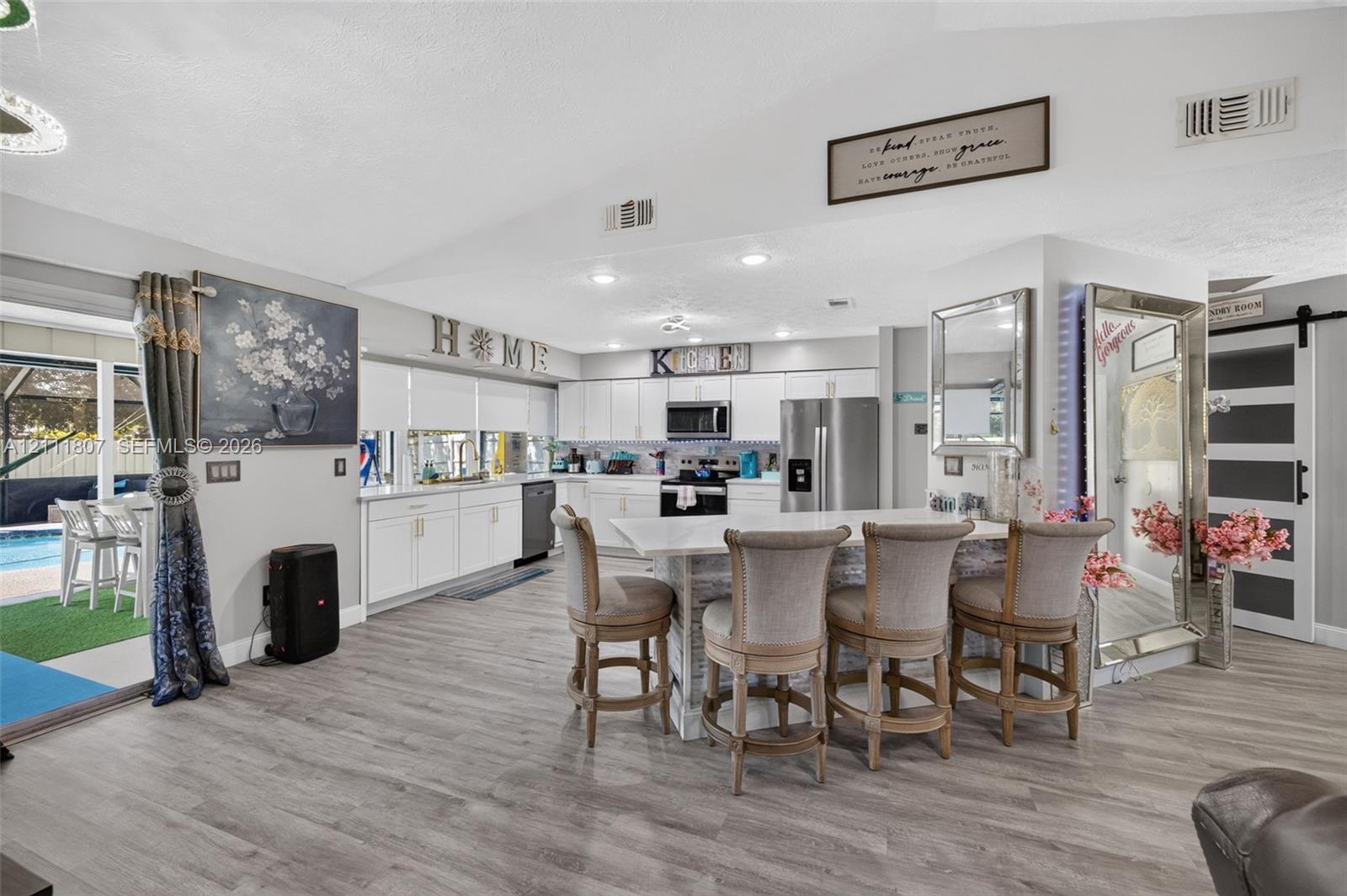 Open-concept kitchen featuring white shaker-style cabinetry, stainless steel appliances, stone-surface central island with bar seating, and wood-finish flooring