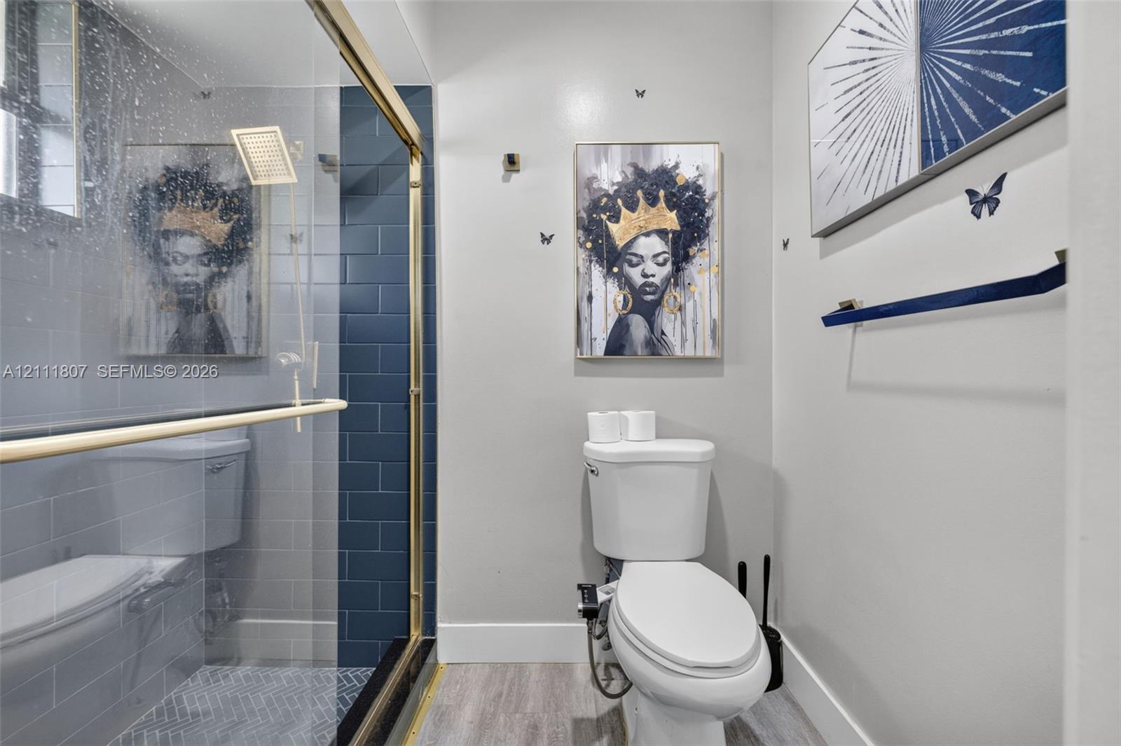 Shower enclosure featuring dark-toned subway tile, a gold-finish frame, and a rainfall showerhead, complemented by wood-finish flooring and a wall-mounted towel bar