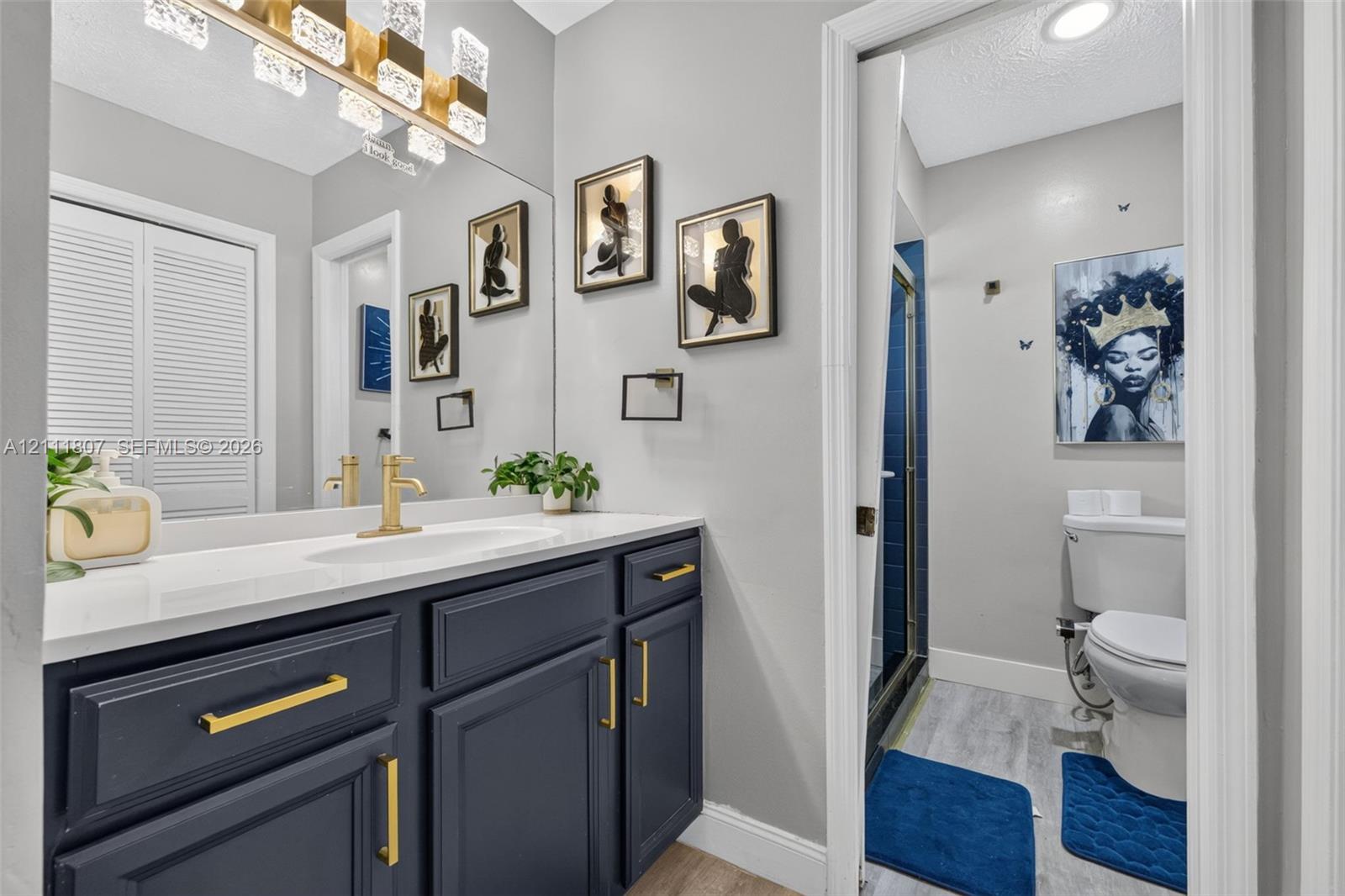 Navy blue vanity with brass-finish hardware, white countertop with integrated sink, brass-finish faucet, rectangular wall-mounted light fixture, and walk-in shower with blue tile surround