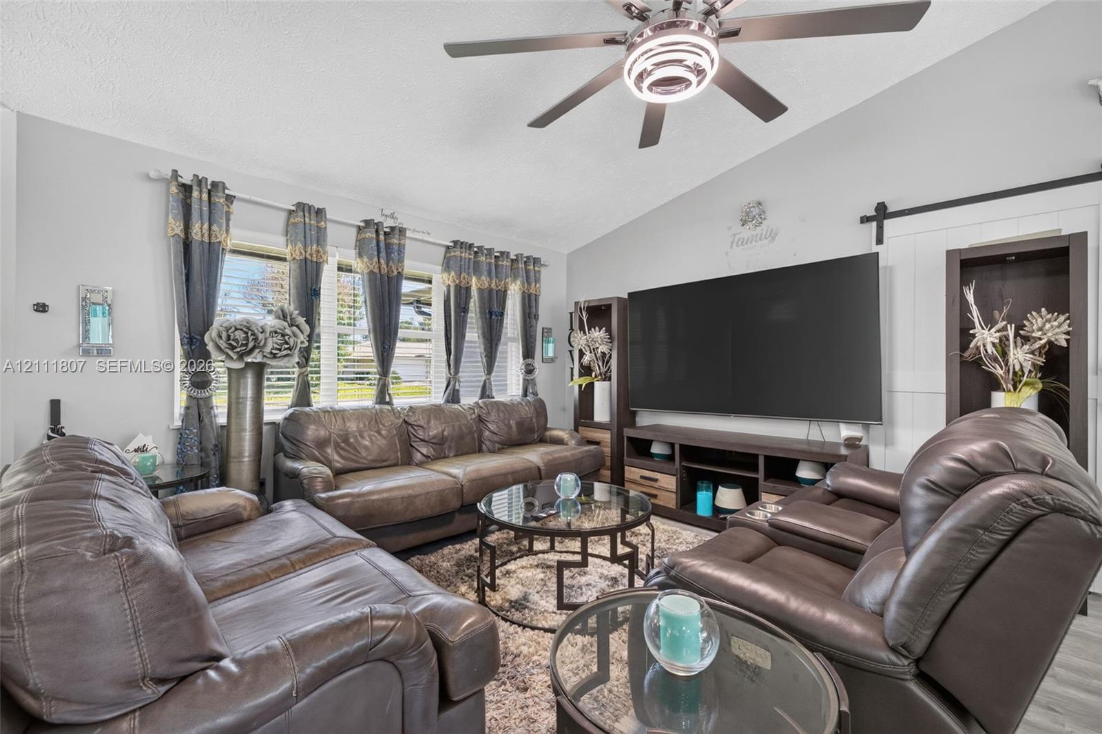 Vaulted ceiling featuring a central multi-blade ceiling fan with integrated lighting, oversized window configuration, and white sliding barn door Vaulted ceiling featuring a central multi-blade ceiling fan with integrated lighting, oversized window configuration, and white sliding barn door